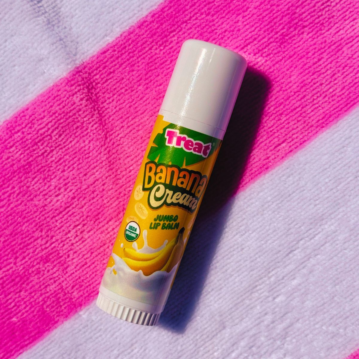 A tube of Treat Banana Cream Jumbo Lip Balm resting diagonally on a textured pink and white striped fabric background.