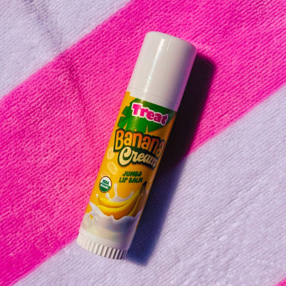 A tube of Treat Banana Cream Jumbo Lip Balm resting diagonally on a textured pink and white striped fabric background.